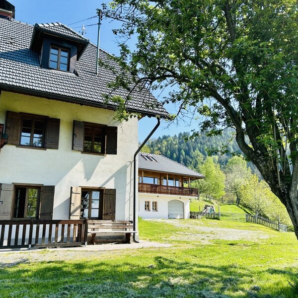 The vacation rental features a multi-story building with a wooden balcony, shutters, a front lawn with a bench, and a view of a green hillside.