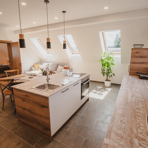 Die voll ausgestattete Küche des Apartments mit Kochinsel inklusive Essbereich und Kaffeemaschine, offen verbunden mit dem Wohnbereich, Sofa und Dachfenstern.