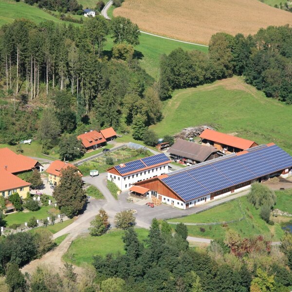 Aerial view of the farmhouse property, including multiple buildings with solar panels, surrounded by forests and green fields.