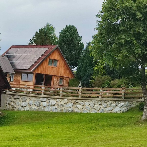 Die Alm ist ein Holzhaus mit Solarpaneelen auf dem Dach, umgeben von grünen Wiesen und Bäumen, mit einer Steinmauer und einem Holzzaun.