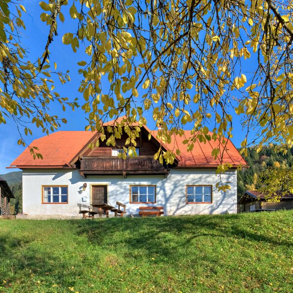 The white vacation rental with a red roof offers a balcony and an outdoor dining area on the lawn, framed by autumn leaves and mountain views.