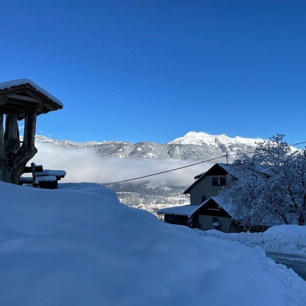 Der verschneite Bauernhof in der Winterlandschaft mit einer rustikalen Holzüberdachung und der Bergkulisse im Hintergrund.