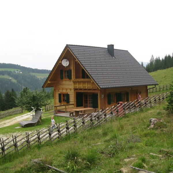 The wooden Alpine Hut features a balcony, an outdoor dining area, and a sun lounger, surrounded by green hills and a wooden fence.