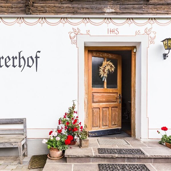 Der Eingangsbereich des Bauernhofs Jöhrerhof mit einer Holztür, dem Hofnamen und der Hausnummer N°4 an der Fassade, Blumen in Töpfen und einer Holzbank.
