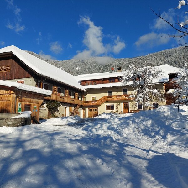 Die Außenansicht des Bauernhofs mit schneebedeckten Dächern und umliegenden Bergen an einem klaren Wintertag.