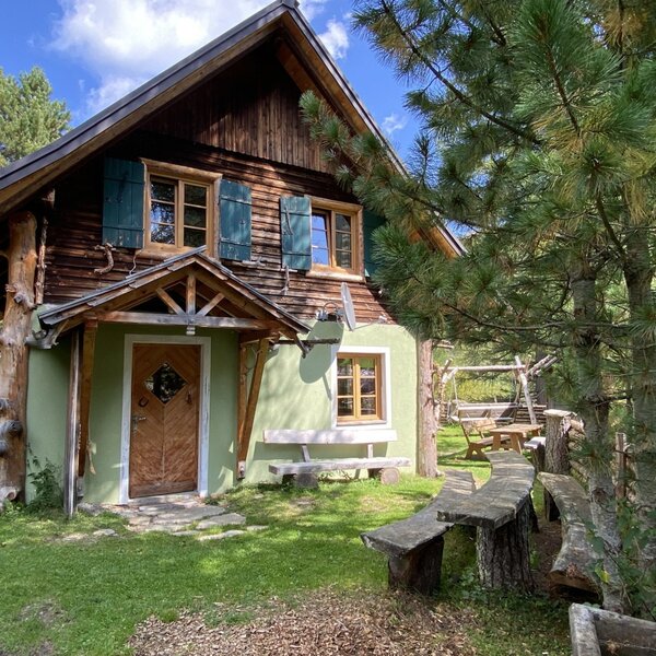 Die Alm von außen mit Holzfassade, grünen Wänden und einem Außenbereich mit Holzbänken und einem Tisch.