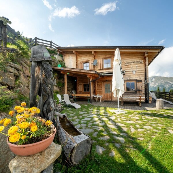 The Alpine Hut features a wooden terrace with outdoor seating, a lawn with stone pathways, and panoramic mountain views.