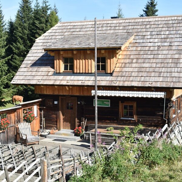 The Alpine Hut features a wooden log exterior, a shingle roof with dormer windows, and a front terrace with seating, surrounded by pine trees, with a cow on an adjacent structure.