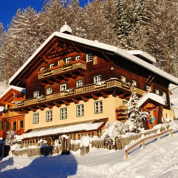 Der Bauernhof im Winter, umgeben von Schnee und Bäumen, mit Holzbalkonen und schneebedecktem Dach.
