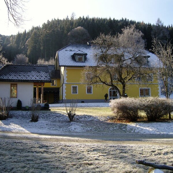 The farmhouse exterior in winter, featuring a yellow main building, a white annex with a dark roof, and surrounding frosty grounds with a forest background.