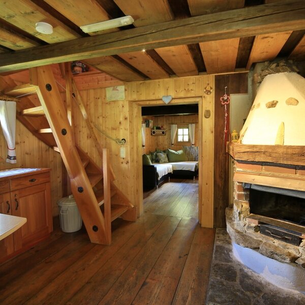 The Alpine Hut's open-plan living area features a wood-fired stove, a wooden staircase leading to the upper floor, and an adjacent kitchen counter.