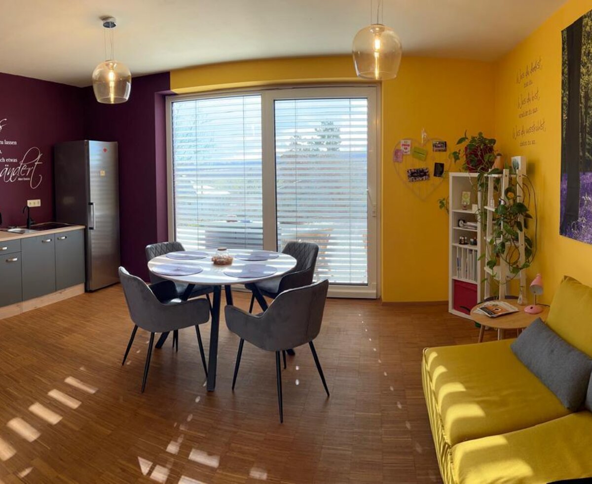 Der kombinierte Wohn- und Essbereich des Appartements verfügt über eine Küchenzeile, einen Esstisch, ein Sofa und Zugang zu einem Balkon.