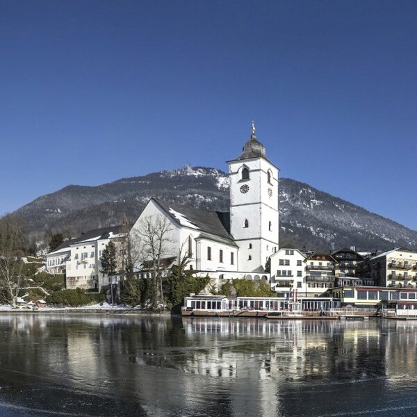 Der Wolfgangsee im Winter mit der Wallfahrtskirche St. Wolfgang und den schneebedeckten Bergen des Salzkammergutes.