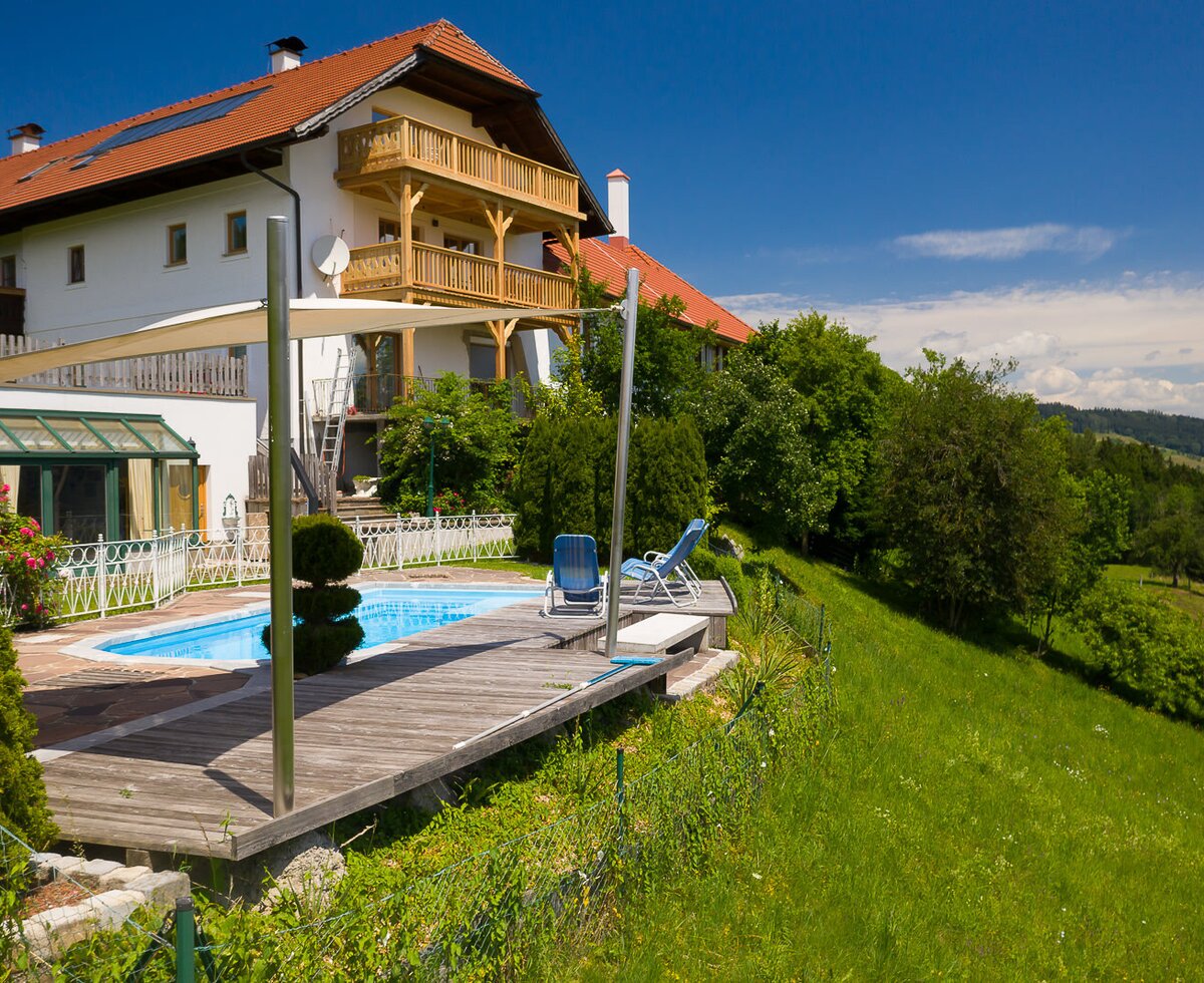 Der Bauernhof mit Außenpool, Holzdeck, Sonnensegel und Blick auf die umliegende grüne Hügellandschaft.