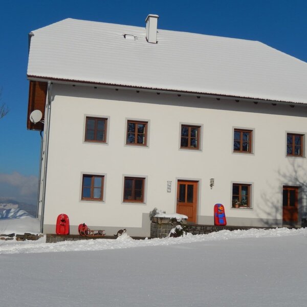 Der zweistöckige Bauernhof mit weißer Fassade, braunen Fenstern und schneebedecktem Dach, umgeben von Winterlandschaft mit Rodeln am Eingang.