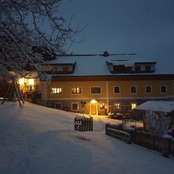 The farm house exterior at night, covered in snow with illuminated windows and a children's swing set.