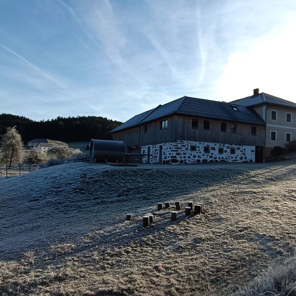 The Farm House exterior features a combination of wood siding, painted walls, and a stone foundation, set on a frost-covered hillside with a large wooden barrel next to the building.