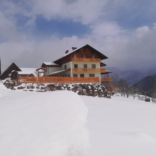Der Bauernhof im Winter, mit Balkonen und einem Holzzaun, umgeben von Schnee und Bergen im Hintergrund.