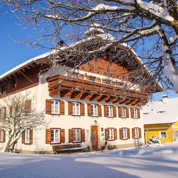 Die winterliche Außenansicht des Bauernhofs mit schneebedecktem Dach, Holzbalkonen und Fenstern mit Holzläden.