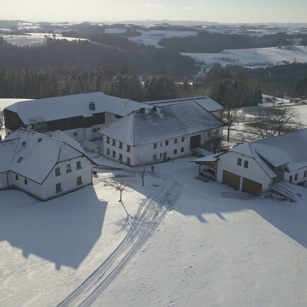The farm house buildings are covered in snow, surrounded by white fields and distant wooded hills in a winter landscape.