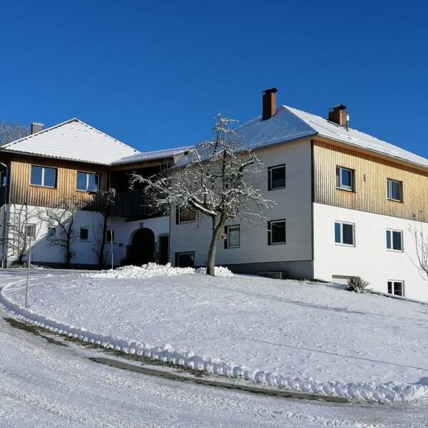 Außenansicht des Bauernhofs im Winter, mit schneebedeckten Dächern und einer Zufahrt unter klarem blauem Himmel.