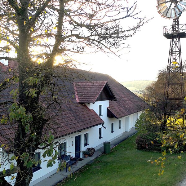 Der Bauernhof mit weißer Fassade, rotem Ziegeldach, umgebender Rasenfläche und einem Windrad.
