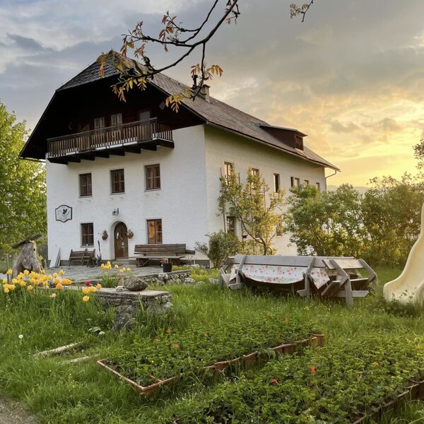 The farmhouse with a balcony, surrounded by a garden with a slide and outdoor seating at sunset.