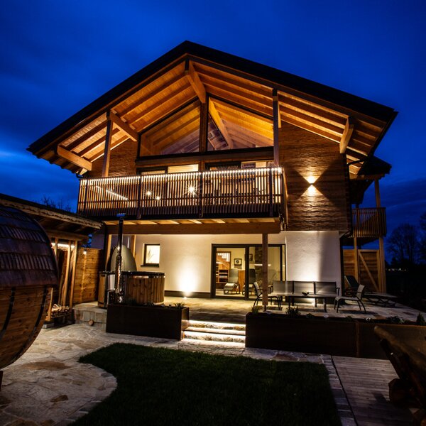 The illuminated Farm House at night, featuring wooden architecture, a balcony, and a ground-floor patio with outdoor seating, a hot tub and a barrel sauna.