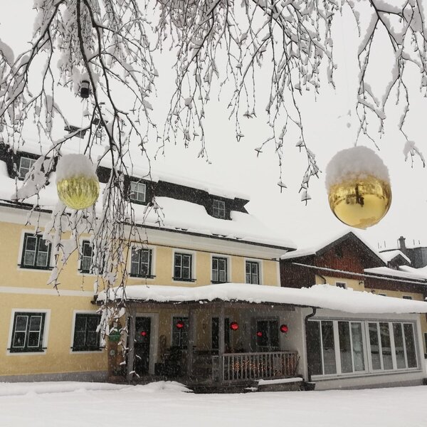 The farm house exterior with snow-covered roofs and trees, featuring a covered porch, a sunroom, and large golden ornaments.