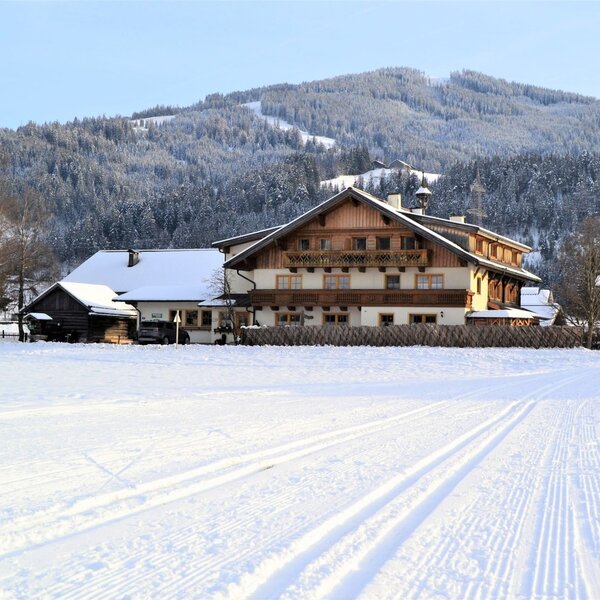 Der Bauernhof Arnoldgut im Winter, umgeben von Schnee und Bergen, mit Langlaufloipen im Vordergrund.