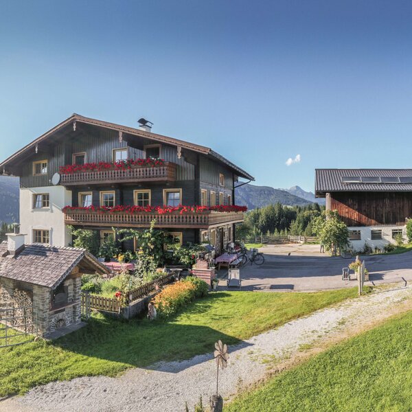 Der Bauernhof mit dem Haupthaus, Balkonen mit Blumen, einem Wirtschaftsgebäude, Kinderspielplatz und umliegenden Wiesen mit Bergblick.