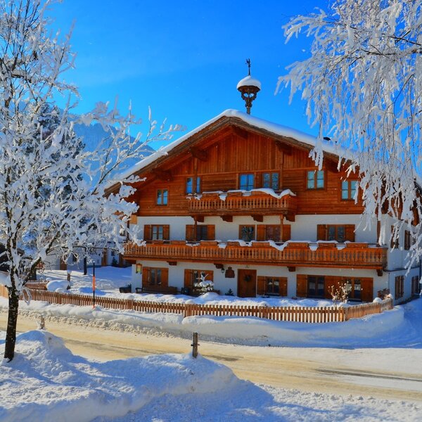 Der Bauernhof im Winter, mit verschneitem Dach, Holzbalkonen und frostbedeckten Bäumen vor blauem Himmel.