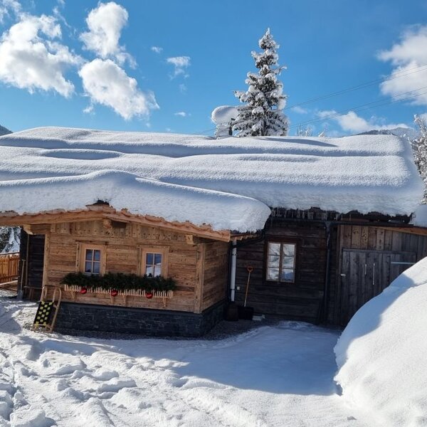 The exterior of the farmhouse, featuring traditional wooden construction heavily covered in snow, set against a backdrop of winter mountains.