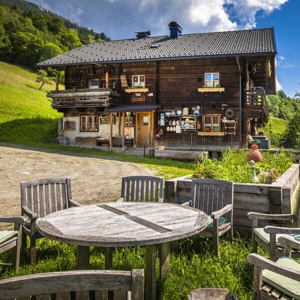Outdoor wooden dining table and chairs with cushions on a grassy area, set in front of the traditional wooden vacation rental on a green hillside.