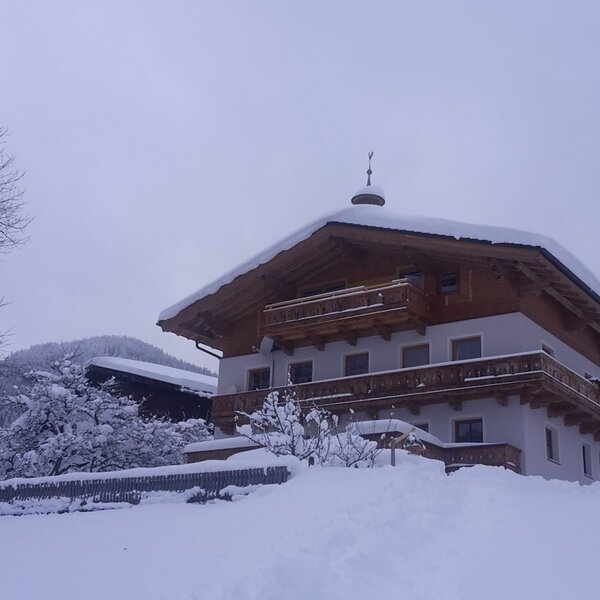 The farm house exterior with traditional wooden balconies and a snow-covered roof, surrounded by snow-covered trees and mountains.