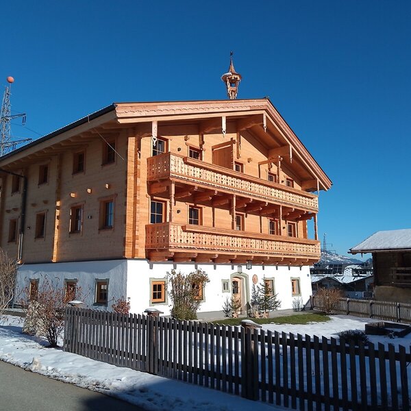 Die Außenansicht des Bauernhofs mit Holzfassade, mehreren Balkonen, einem Glockenturm auf dem Dach und einem weiß verputzten Erdgeschoss, umgeben von einem Gartenzaun und Schnee.