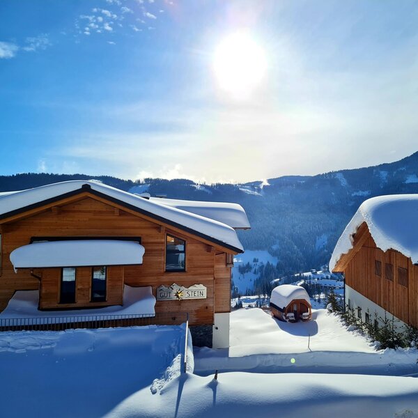 The farm house "GUT STEIN" and adjacent building, both snow-covered, feature a barrel-shaped outdoor structure with a mountain landscape in the background.