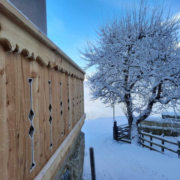 Das frostbedeckte Holzbalkongeländer des Bauernhofs mit traditionellen Mustern, umgeben von einer verschneiten Winterlandschaft mit Baum und Zaun.