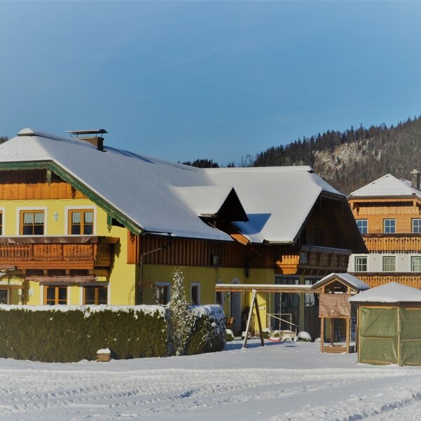 The Farm House exterior with a snow-covered roof, wooden balcony, and a swing set in the snowy yard.