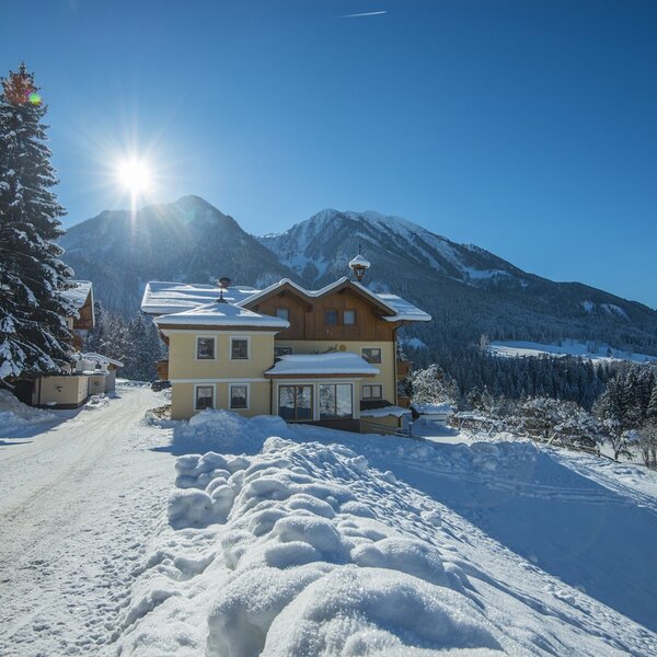 Der Bauernhof im Winter, umgeben von Schnee und Bergen, bei Sonnenschein.