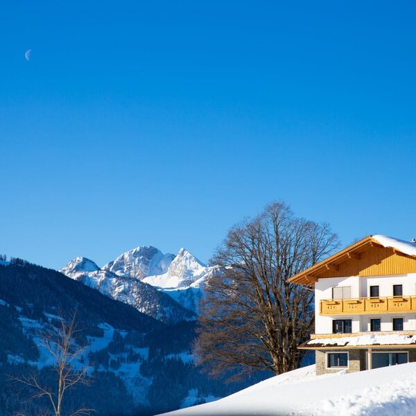 Der Bauernhof im Winter, mit schneebedecktem Dach und Holzbalkonen, vor der Kulisse der verschneiten Salzburger Bergwelt unter klarem blauem Himmel.