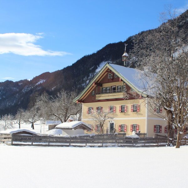 Der Bauernhof mit gelber Fassade, roten Fensterläden und schneebedecktem Dach in winterlicher Landschaft mit Bergen.
