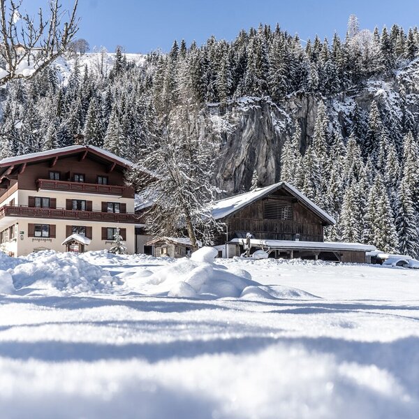 Der Bauernhof mit seinen Gebäuden in einer tief verschneiten Winterlandschaft vor Nadelwäldern und einer Felswand.
