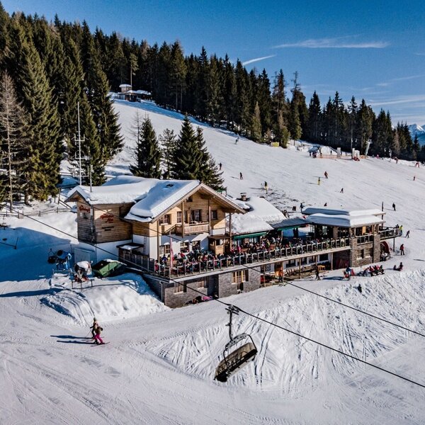 The farmhouse, a multi-story wooden building, is situated directly on the ski slope, featuring a large outdoor terrace for guests.