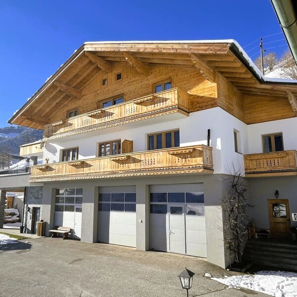 Außenansicht des Bauernhofs mit Holzbalkonen, weißer Fassade und Garagentoren, umgeben von Schnee.