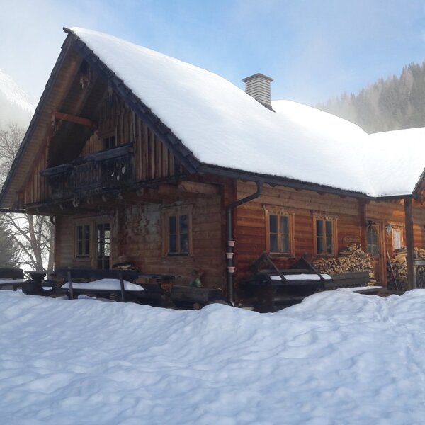 The wooden farmhouse exterior, featuring a snow-covered roof, a balcony, and stacked firewood on the porch.