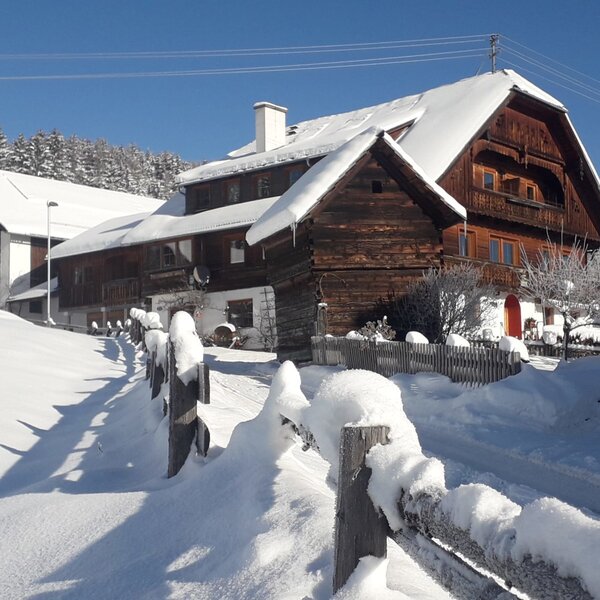 Außenansicht des Bauernhofs im Winter, mit verschneitem Dach, einem Holzzaun und Nadelbäumen im Hintergrund.