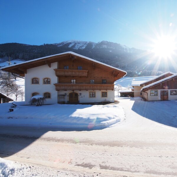 Die Außenansicht des Bauernhofs in einer verschneiten Berglandschaft bei Sonnenschein.