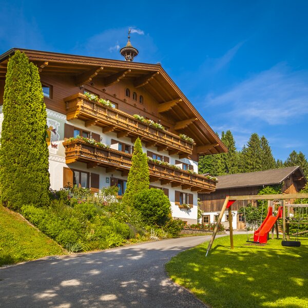 Der Bauernhof mit blumengeschmückten Balkonen und einem Kinderspielplatz auf der Rasenfläche.