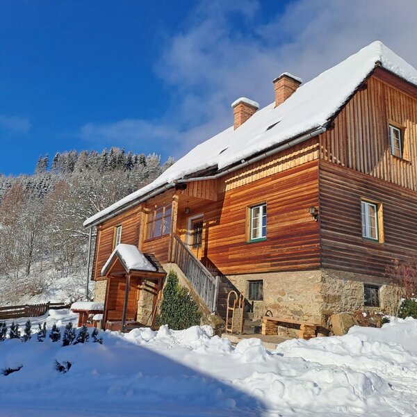 The snow-covered farmhouse, showcasing its wooden exterior, stone base, and an outdoor oven in a winter setting.